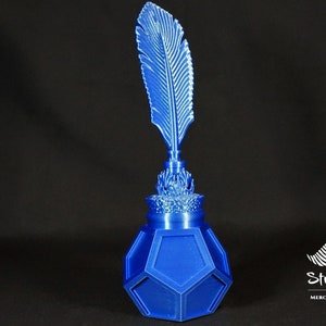 May include: A blue inkwell with a feather quill design. The inkwell has a geometric shape and a decorative top. The feather quill is upright, and the entire piece is a vibrant blue color. The Stratation design logo is visible.