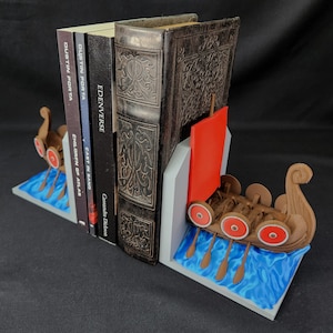 May include: A set of Viking ship bookends. The bookends are brown with red and white accents, and feature a ship design with oars and shields. The base is blue and white, resembling water. Books are held between the bookends.