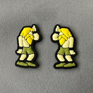 Puede incluir: Dos dijes de zapatos estilo caricatura. Cada dije presenta una figura con cabello amarillo, barba blanca y pantalones cortos verdes. Las figuras están delineadas en negro y parecen estar en una pose con una mano levantada. Los dijes están sobre un fondo gris.