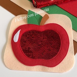 Puede incluir: Una manzana de fieltro roja con un centro de lentejuelas rojas. La manzana está sobre un fondo de fieltro beige. El texto "Feltro con fantasia" es visible en la imagen. El texto "PDF y video tutorial" también es visible en la imagen.