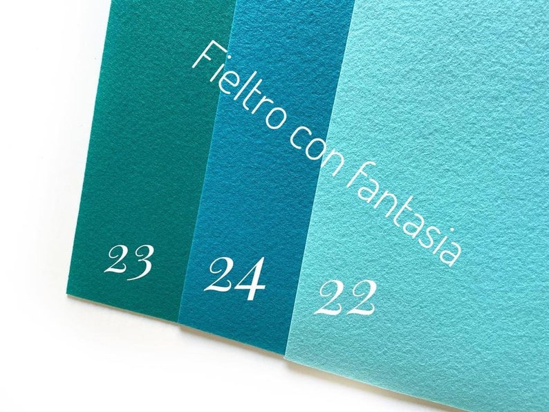 May include: Three felt fabric swatches in shades of teal and turquoise. The swatches are labeled with the numbers 23, 24, and 22. The text "Feltro con fantasia" is written diagonally across the swatches.