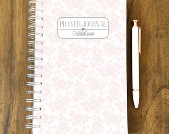 May include: A spiral-bound prayer journal with a floral pattern in shades of pink and white. The cover features the text "PRAYER JOURNAL" and the name "Addison". A white pen with gold accents rests beside the journal on a wooden surface.