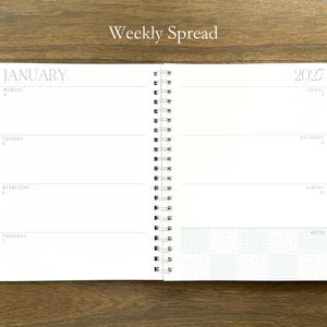 Puede incluir: Una agenda de espiral abierta en una vista semanal para enero de 2027. Las p&aacute;ginas blancas presentan texto en negro que incluye el encabezado Weekly Spread, el mes de enero, el a&ntilde;o 2027 y los d&iacute;as de la semana con una secci&oacute;n de notas.