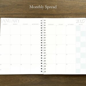 Puede incluir: Una agenda de espiral abierta en una vista mensual para enero de 2027. Las p&aacute;ginas blancas presentan una cuadr&iacute;cula con los d&iacute;as de la semana, fechas numeradas y una columna de notas con un patr&oacute;n de cuadros. El texto Monthly Spread es visible.