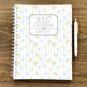 May include: A spiral-bound 2027 calendar personalized with the name KRISTEN, featuring a repeating pattern of yellow lemons and light blue bows on a white background with vertical light blue stripes, accompanied by a white pen with gold accents.