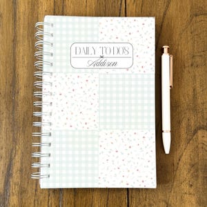 May include: A spiral-bound notebook with a cover featuring a patchwork design of pastel green gingham and floral patterns. The notebook has the text "DAILY TO DO'S" and the name "Addison". A white pen with rose gold accents rests beside the notebook.