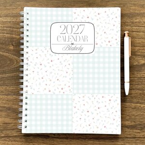 Puede incluir: Un calendario planificador 2027 de espiral titulado Blakely con un dise&ntilde;o de portada de patchwork que alterna cuadros vichy azul claro y un peque&ntilde;o patr&oacute;n floral en rosa y azul, acompa&ntilde;ado de un bol&iacute;grafo blanco con detalles en oro rosa.