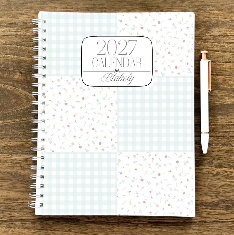 Puede incluir: Un calendario planificador 2027 de espiral titulado Blakely con un dise&ntilde;o de portada de patchwork que alterna cuadros vichy azul claro y un peque&ntilde;o patr&oacute;n floral en rosa y azul, acompa&ntilde;ado de un bol&iacute;grafo blanco con detalles en oro rosa.