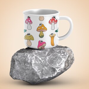 May include: White ceramic mug with a colorful cartoon mushroom pattern. The mug has a handle and is sitting on a gray rock.