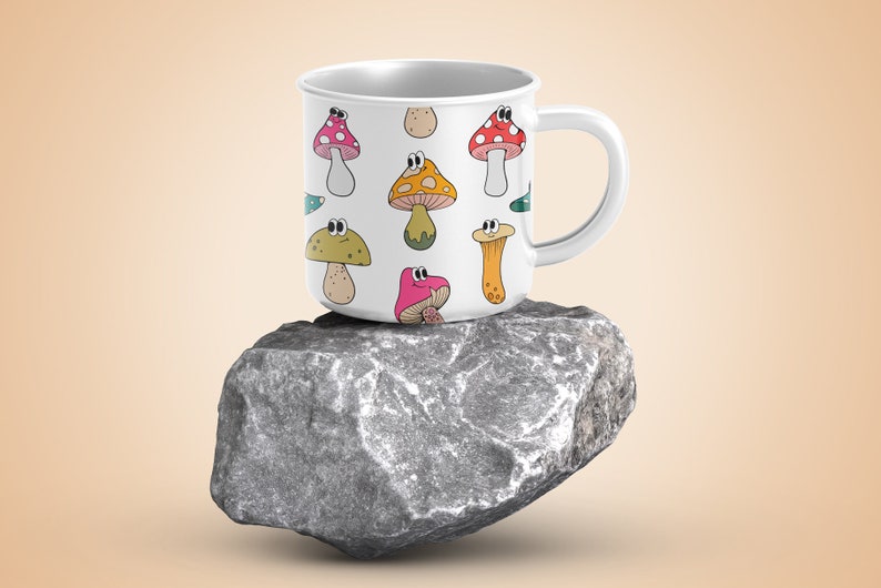 May include: White ceramic mug with a colorful cartoon mushroom pattern. The mug has a handle and is sitting on a gray rock.
