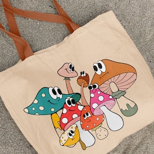 May include: A beige canvas tote bag with a colorful cartoon design of mushrooms with googly eyes. The mushrooms are various colors, including red, orange, green, and yellow.