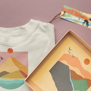 May include: A white t-shirt with a graphic of a mountain range, a sun, and birds flying. The graphic is in shades of pink, yellow, orange, brown, and blue. The t-shirt is next to a brown box with a similar graphic inside.