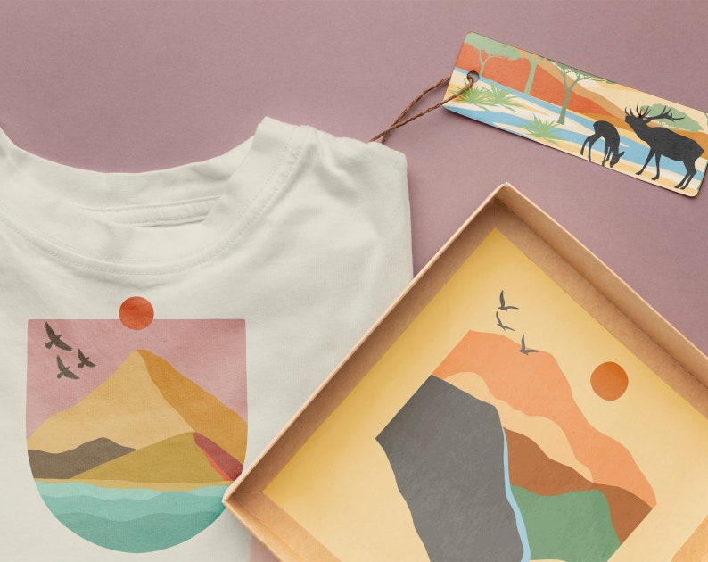 May include: A white t-shirt with a graphic of a mountain range, a sun, and birds flying. The graphic is in shades of pink, yellow, orange, brown, and blue. The t-shirt is next to a brown box with a similar graphic inside.