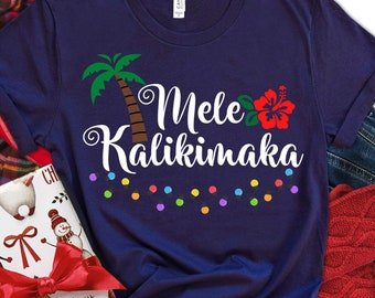 May include: A navy blue t-shirt featuring a green palm tree, a red hibiscus flower, and the white text Mele Kalikimaka above a row of colorful dots. The shirt is displayed with a red plaid scarf, blue jeans, and a gift box with Christmas graphics.