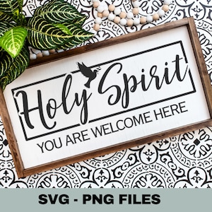 May include: A rectangular wooden sign with the words "Holy Spirit" in black script, a dove graphic, and the phrase "You Are Welcome Here." The sign has a white background and a dark brown frame. Greenery and beads are also visible.