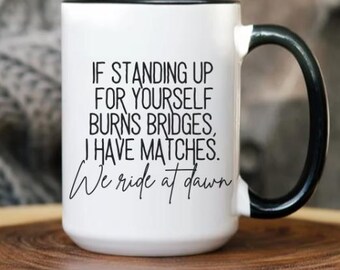 May include: White ceramic mug with a black handle and rim. The mug features the text "IF STANDING UP FOR YOURSELF BURNS BRIDGES, I HAVE MATCHES. We ride at dawn" in black lettering. The mug is on a wooden surface.