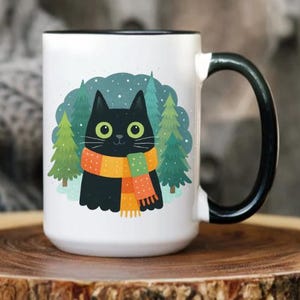 May include: White ceramic mug with a black handle and rim, featuring a cartoon black cat wearing an orange and yellow scarf. The cat is set against a backdrop of green trees and falling snow. The mug is on a wooden surface.