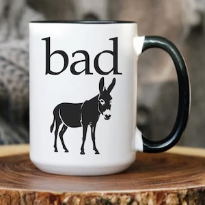 May include: White ceramic mug with a black handle and rim. The mug features the word "bad" in black capital letters above a black silhouette of a donkey. The mug is on a wooden surface.