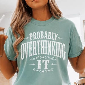 May include: A seafoam green short-sleeve t-shirt featuring white text that reads "PROBABLY OVERTHINKING IT" in a decorative font. The shirt has a crew neckline and a relaxed fit, paired with white shorts that have black stripes on the cuffs.