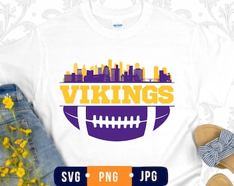 May include: A white short-sleeve t-shirt featuring a graphic print of a city skyline in yellow and purple above the word VIKINGS in bold yellow letters, with a purple and white football illustration positioned below the text on the front.