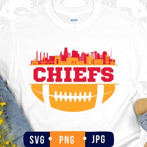 May include: A white t-shirt featuring a graphic design with a red and orange city skyline silhouette above the word CHIEFS in bold red capital letters, positioned over a stylized orange football illustration with white laces and stripes.