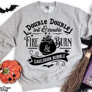 Pu&ograve; includere: Felpa grigia con un design grafico nero che presenta un calderone, pipistrelli e il testo "Double Double toil & trouble Fire Burn & Cauldron Bubble".