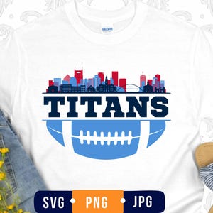 May include: A white t-shirt featuring a graphic design with a city skyline in red, light blue, and dark blue above the word TITANS in bold dark blue letters, followed by a light blue football illustration with white laces and a white outline.