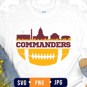 May include: A white t-shirt featuring a maroon graphic of the Washington D.C. skyline above the word COMMANDERS in maroon text, with a yellow football graphic below. The shirt has a Gildan brand label at the neck and a crew neckline design.
