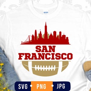 May include: A white short-sleeve t-shirt featuring a red graphic of the San Francisco skyline, including the Golden Gate Bridge, above the red text SAN FRANCISCO and a gold football illustration. The shirt has a crew neckline and short sleeves.