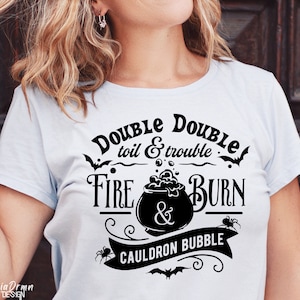 Pu&ograve; includere: Una maglietta blu chiaro con un design grafico nero che presenta un calderone, pipistrelli e ragni. Il testo sulla maglietta recita "Double Double toil & trouble Fire & Burn Cauldron Bubble".