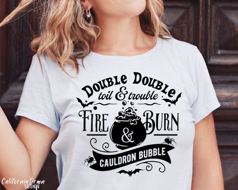 Pu&ograve; includere: Una maglietta blu chiaro con un design grafico nero che presenta un calderone, pipistrelli e ragni. Il testo sulla maglietta recita "Double Double toil & trouble Fire & Burn Cauldron Bubble".