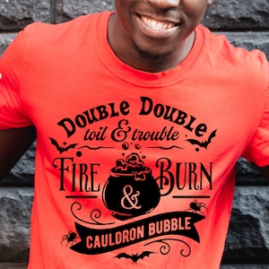 Pu&ograve; includere: Una maglietta rossa con un design grafico nero che dice "Double toil & trouble Fire & Burn Cauldron Bubble". La grafica include un calderone con bolle, pipistrelli e ragni.
