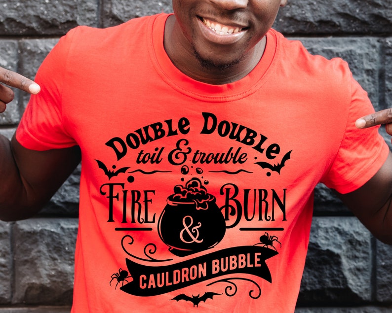 Pu&ograve; includere: Una maglietta rossa con un design grafico nero che dice "Double toil & trouble Fire & Burn Cauldron Bubble". La grafica include un calderone con bolle, pipistrelli e ragni.