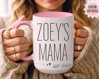 May include: White ceramic mug with a pink interior and handle. The mug features the text "ZOEY'S MAMA" in black, with "est. 2021" below. The mug is held by a person wearing a beige sweater.
