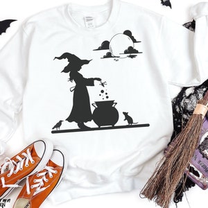 May include: White sweatshirt with a black silhouette of a witch stirring a cauldron under a full moon. A crow and a mouse are also in the design. Orange sneakers are in the foreground. The words "California Dream Design" are at the bottom.