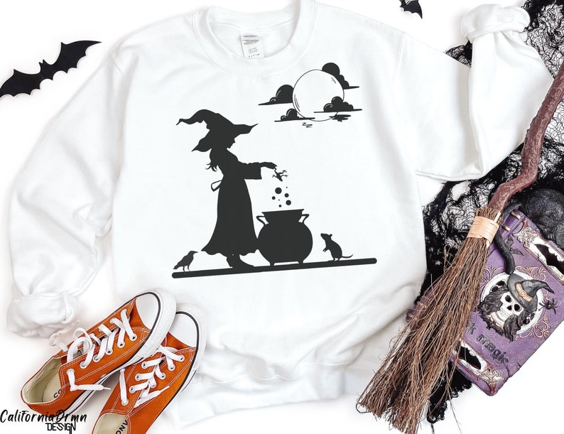 May include: White sweatshirt with a black silhouette of a witch stirring a cauldron under a full moon. A crow and a mouse are also in the design. Orange sneakers are in the foreground. The words "California Dream Design" are at the bottom.
