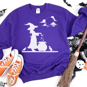 May include: A purple sweatshirt with a white graphic of a witch stirring a cauldron under a moonlit sky. The design includes a witch, a cauldron, a mouse, and a crow. The sweatshirt is paired with orange sneakers.