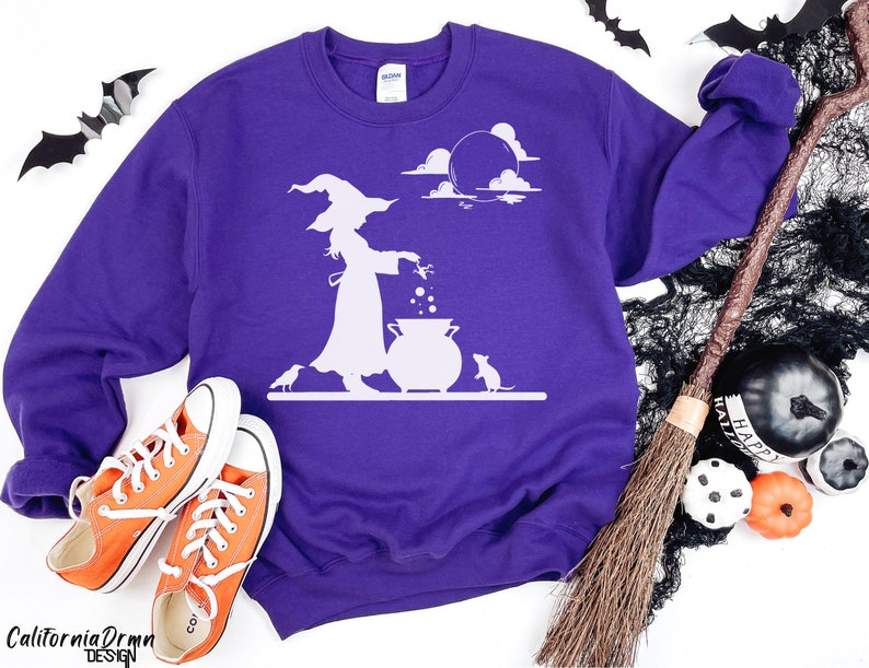May include: A purple sweatshirt with a white graphic of a witch stirring a cauldron under a moonlit sky. The design includes a witch, a cauldron, a mouse, and a crow. The sweatshirt is paired with orange sneakers.