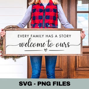 May include: A wooden framed sign with the text "EVERY FAMILY HAS A STORY welcome to ours" in black script. The sign is held by a person wearing a red and blue plaid vest and blue jeans. A wreath is attached to the left side of the sign.