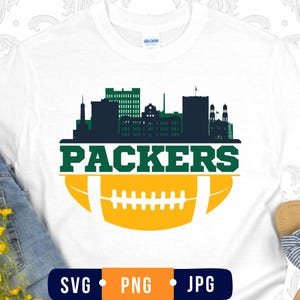 May include: A white t-shirt featuring a graphic design with a dark green city skyline silhouette above the word PACKERS in bold green letters, positioned over a yellow football illustration with white laces and stripes on a white background.