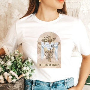 May include: White t-shirt with a graphic of a cross with a white cloth draped over it, surrounded by leaves and a marble background. The text "HE IS RISEN" is printed below the graphic.