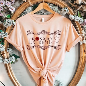 May include: A light pink t-shirt with a graphic design of a red rose and the text "Rosary Extremist" in a dark red font.