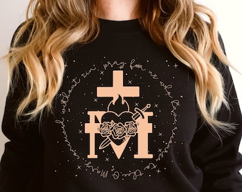 May include: Black sweatshirt with a peach-colored graphic of a heart with a sword and roses, surrounded by a circle of text that reads "Mother, I pray for us who are without sin."