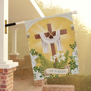 Könnte beinhalten: Dekorative Outdoor-Flagge mit einem braunen Kreuz und weißem Tuch vor einem gelben Hintergrund mit Sonnenstrahlen. Weiße Lilien und der Text "HE IS RISEN" sind ebenfalls abgebildet.