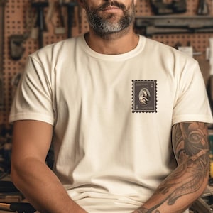 Könnte beinhalten: Ein weißes T-Shirt mit einem Vintage-Stempeldesign, das ein Schwarzweißbild einer Frau und eines Kindes zeigt. Das Shirt wird von einer Person mit einem Tattoo am Arm getragen.