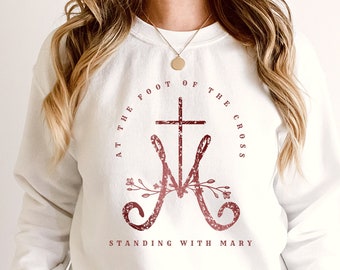 May include: White sweatshirt with a maroon graphic design featuring a cross and the text "At the foot of the cross, standing with Mary".