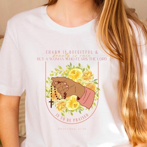 May include: White t-shirt featuring a floral design and a hand holding a rosary. The text on the shirt reads "Charm is deceitful & beauty is vain, but a woman who fears the Lord is to be praised, Proverbs 31:30."