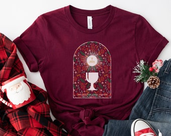 Op de afbeelding: Bordeauxrood T-shirt met een afbeelding van een kelk en de tekst "Remember the Lord's Supper". De afbeelding bevat een blauwe jeans, rode sneakers, een rood-zwarte geruite sjaal en een kerstmanmok.