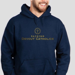 Pode incluir: Camisola de capuz azul marinho com um gráfico dourado de uma cruz dentro de um círculo com as palavras "Raising Devout Catholics" abaixo.