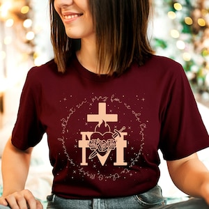 May include: A maroon t-shirt with a gold design featuring a heart with a cross, a sword, and roses. The text around the design reads "With Christ I can do all things. Pray for us who labor in the vineyard."
